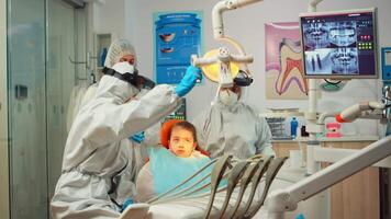 Dentistry doctor in protective suit using sterilized dental tools examining kid patient during coronavirus pandemic. Medical team talking with woman wearing face shield, coverall, mask and gloves video