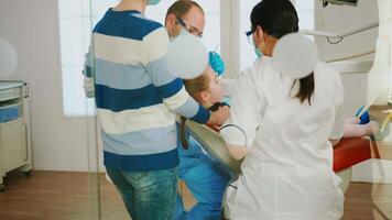 odontología doctores limpieza dientes de niño, tratando dental problemas mientras madre en pie cerca estomatológico silla hablando con muchacha. dentista elimina diente decaer perforación dientes durante cirugía video