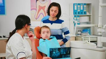 pediátrico dentista demostración dental problemas participación radiografía señalando en afectado dientes mientras hombre asistente preparando esterilizado herramientas para cirugía. médico y enfermero trabajando en estomatológico unidad video