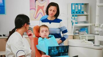 Pediatric dentist showing dental problems holding radiography pointing on affected teeth while man assistant preparing sterilized tools for surgery. Doctor and nurse working in stomatological unit video