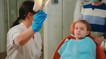 femme stomatologue technicien éclairage le lampe pour examiner peu patient séance sur stomatologique chaise. médecin Parlant à fille vérification les dents santé tandis que infirmière en train de préparer outils pour examen video