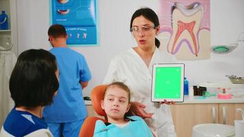 Stomatologist woman looking at green screen tablet while speaking with mother of little patient. Pediatric dentist explaining using monitor with green chroma key izolated chroma pc key mockup display video