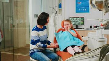 menina mostrando para mãe afetado massa sentado em estomatológico cadeira falando com dela mãe enquanto equipe do estomatologista médicos analisando dentes raio X do pequeno paciente com dor de dente dentro fundo video