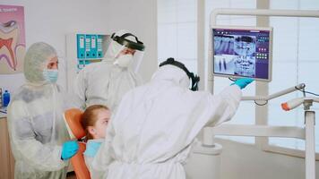estomatólogo en bata explicando dental problema utilizando digital monitor durante global pandemia. asistente y médico con bata cara proteger máscara guantes examinando mujer desde estomatológico silla video