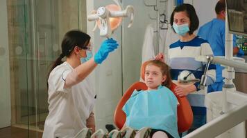 Pediatric dentist and assistant treating girl patient in modern stomatologic clinic. Nurse and doctor wearing protection masks cleaning teeth of child in dental office lying on chair with open mouth video