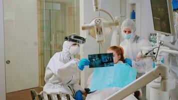 Doctor in protective suit discussing about x-ray of tooth with mother of child patient explaining treatment using tablet in covisd-19 pandemic. Medical team wearing face shield, coverall, mask, gloves video