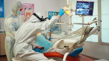 dentaire technicien dans protecteur équipement éclairage le lampe pour examiner enfant patient pendant covid-19 épidémie. médical équipe parlant avec mère portant visage bouclier, combinaison costume, masque et gants video