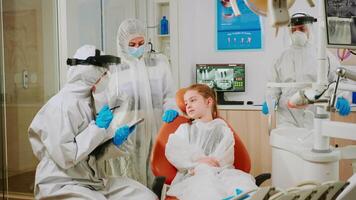 dentista medico nel tuta parlando con ragazza paziente durante globale coronavirus pandemia scrittura su clipboard. infermiera e ortodontico medico indossare viso scudo, protezione completo da uomo, maschera e guanti. video