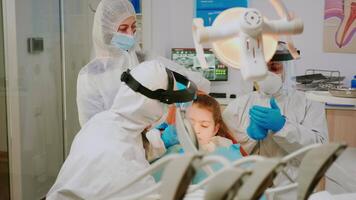 Orthodontist doctor in protection suit drilling kid tooth in dental clinic during coronavirus pandemic. Nurse and doctor wearing face shield, protection suit, mask and gloves talking with patient video