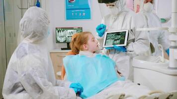 stomatolog i skyddande Utrustning som visar på läsplatta dental röntgen granskning den med mor av patient. medicinsk team bär ansikte skydda mask, handskar, förklara radiografi använder sig av anteckningsbok visa video