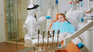 Dentist with coverall holding mouth x-ray image child patient talking with patient mother during global pandemic. Assistant and doctor talking wearing suit, coverall, protection suit, mask, gloves video