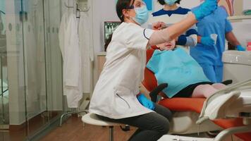 femme dentiste technicien éclairage le lampe pour examiner peu patient. orthodontiste Parlant à fille séance sur stomatologique chaise tandis que infirmière en train de préparer outils pour examen dans moderne clinique. video