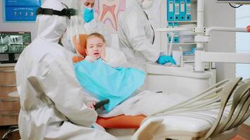 criança vestindo proteção terno usando dedo para ponto afetado dente enquanto dentista dentro macacão falando com mãe antes estomatológico exame durante covid-19 pandemia dentro Novo normal dental clínica video