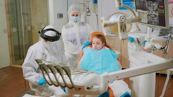 Child with ppe suit taking care of dental health during coronavirus pandemic. Orthodontist doctor lighting the lamp wearing face shield, protection suit, mask and gloves talking with patient video