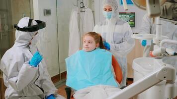 pequeno menina com dental babador e ppe terno ouvindo pediatra dentista dentro macacão sentado dentro dental cadeira durante coronavírus pandemia. estomatologista vestindo proteção terno, face escudo mascarar luvas video