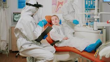 pediátrico dentista en ppe traje hablando con niño paciente escritura en portapapeles durante global coronavirus pandemia. enfermero y ortodoncia médico vistiendo cara proteger, proteccion traje, máscara y guantes. video