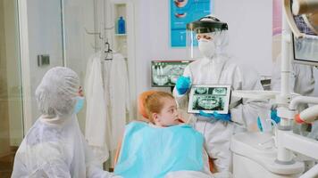 Dentist with face shield explaining panoramic mouth x-ray image to mother of kid patient during global pandemic. Stomatologist talking with woman wearing suit, coverall, protection suit, mask, gloves video