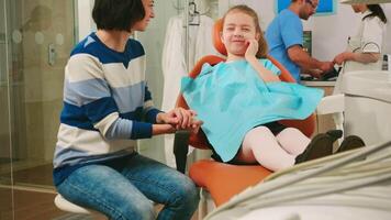 Pediatric dentist checking teeth x-ray of kid patient talking with man assistant in background. sad child showing affected mass to mother sitting stomatological chair dental unit. video