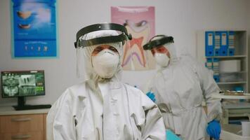 Portrait of tired stomatologist woman with coverall and face shield looking at camera sitting in new normal dental office. pediatric nurse talking with child patient in background. video