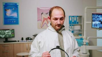 Portrait of exhausted stomatologist man taking off face shield looking at camera sitting in new normal dental office. pediatric nurse talking with child patient in background. video