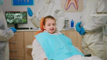 fechar acima do feliz menina paciente rindo às Câmera visitando dentista durante global pandemia, esperando para pediatra estomatologista. médico equipe vestindo ppe terno, face escudo mascarar, luvas examinando criança video