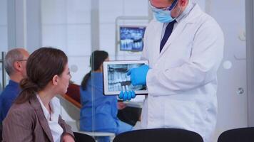 Woman sitting on chair in waiting room of crowded stomatological clinic listening doctor looking on tablet with digital x-ray. Dentist showing to patient teeth radiography using modern gadget. video