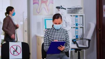 Patient with face protection mask writing on registration form in stomatomoly clinic, sitting in reception respecting social distance. Assistant in coverall measuring patient temperature video