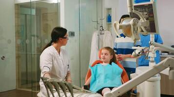 pediatra dentista explicando para criança limpeza processo do dentes enquanto homem assistente preparando esterilizado Ferramentas para exame. enfermeira e médico trabalhando juntos dentro moderno estomatológico clínica video