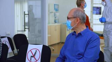 ritratto di anziano paziente con protezione maschera parlando seduta su sedie conservazione sociale distanza nel stomatologiche clinica, in attesa per medico durante coronavirus. concetto di nuovo normale dentista visitare video