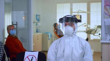 Portrait of tired doctor in dental office looking on camera wearing overall and face shield sitting on chair in waiting room clinic. Concept of new normal dentist visit in coronavirus outbreak. video