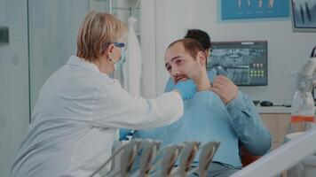 Sénior dentiste examiner patient avec sérieux mal aux dents, avoir prêt à faire stomatologique consultation avec dentaire outils dans armoire. médical traiter homme dans douleur avec oral se soucier problèmes. video