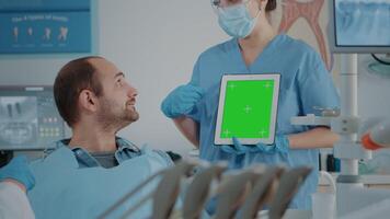 Stomatology nurse vertically holding tablet with green screen, explaining isolated copy space with chroma key background and mock up template to patient at oral care examination. video