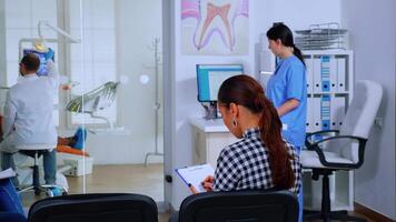 Back view of woman filling in dental document sitting on chiar in waiting room preparing for teeth exemination while doctor working in background. Concept of crowded professional orthodontist office. video