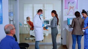 Dentistry doctor showing x-ray of teeth to patient using tablet standing in waiting area of dental clinic . Stomatologist reviewing dental radiography with woman explaining treatment in crowded office video