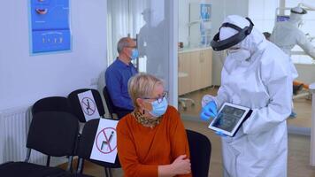 tandarts dokter met gezicht schild richten Aan tablet Scherm uitleggen tandheelkundig röntgenstraal naar senior geduldig gedurende globaal pandemie. verpleegster vervelend bescherming pak, algemeen, masker en handschoenen, nieuw normaal video