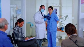 Dentist showing teeth x-ray reviewing it with nusre. Doctor and assistant working in modern crowded stomatological clinic, patients sitting on chairs in reception filling in dental forms and waiting video