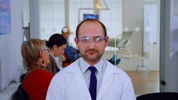 Portrait of stomatologist smiling at camera being in dental office while patients waiting him in background. Dentistry doctor looking on webcam sitting on chair in stomatological clinic. video