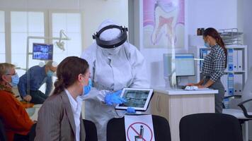 estomatólogo en protector traje señalando en digital radiografía de diente explicando a paciente tratamiento utilizando tableta en covid-19 pandemia. médico equipo vistiendo cara proteger, sobretodo, máscara y guantes. video