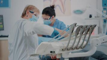 tandheelkunde team onderzoeken geduldig in stomatologie kastje, gebruik makend van tandheelkundig gereedschap naar traktatie kunstgebit problemen. verpleegster en tandarts overleg plegen Mens, aan het doen stomatologisch procedure Bij controle op bezoek komen. video