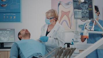 Dentist with face mask using dental tools to examine denture, consulting patient with teeth problems. Stomatology expert doing oral care inspection on man to cure toothache. video