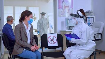 Dentist assistant with ppe equipment talking with patient before consultation during coronavirus epidemic sitting on chairs in waiting area keeping distance. Concept of new normal dentist visit. video