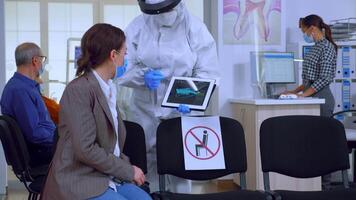 Dentist in coverall using tablet explaining dental x ray to patient in stomatologic office during coronavirus. Nurse wearing face shield, mask showing to woman radiography using digital device video