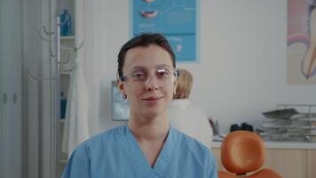Portrait of orthodontic nurse looking at camera in dentistry office. Woman assistant wearing protective glasses before attending stomatological procedure at oral care clinic. Close up video