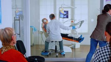 Patients asking informations filling in dental document preparing for teeth exemination. Senior woman sitting on chair in waiting area of crowded orthodontist office while doctor working in background video