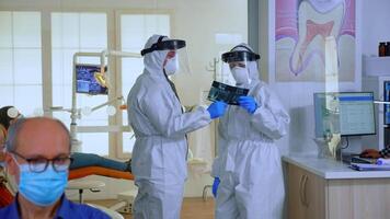 lento movimento di stomatologiche medici con complessivamente guardare a raggi X nel in attesa la zona, pianificazione chirurgia durante globale pandemia mentre anziano pazienti discutere seduta su sedie conservazione distanza. video