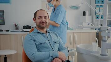 portrait de patient séance dans dentaire chaise à oral se soucier clinique, à la recherche à caméra et souriant. caucasien homme ayant stomatologique rendez-vous avec dentisterie spécialiste dans armoire. video