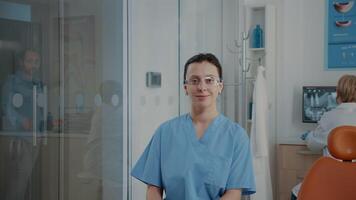 Portrait of dentistry nurse with uniform and protective glasses in stomatology cabinet. Confident assistant preparing for oral care examination, doing dental practice. Woman looking at camera video