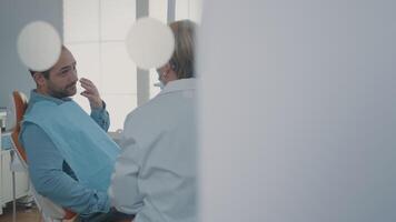 dentiste montrer du doigt à dentier radiographie sur numérique tablette, expliquant diagnostic à patient avec carie. médical montrant X rayon analyse résultats à homme dans douleur à faire chirurgical procédure dans armoire. video