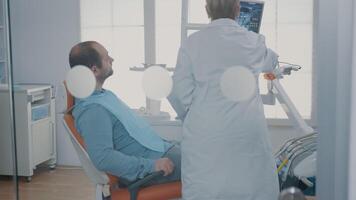 médical montrer du doigt à X rayon analyse diagnostic sur moniteur dans dentisterie armoire, expliquant radiographie résultats à patient dans dentaire chaise. dentiste Faire vérification examen avec homme dans douleur. video