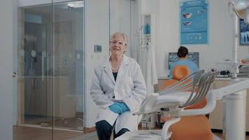 portrait de dentiste avec des lunettes séance dans stomatologie bureau, à la recherche à caméra. dentisterie expert avoir prêt à consulter patient, travail avec dentaire équipement à oral se soucier clinique. video